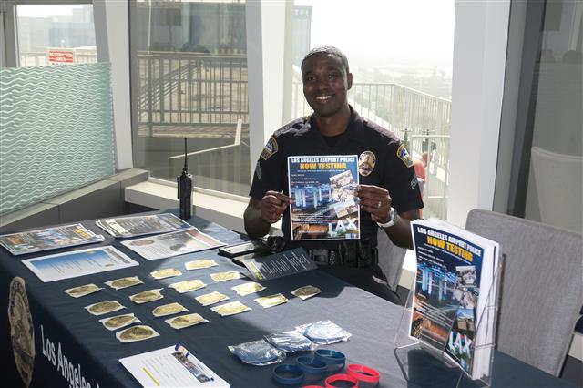 Airport police officer sitting at a Los Angeles Airport Police booth at the event, he is smiling and holding up a flyer. 